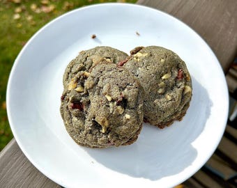 May include: Three cookies on a white plate. The cookies are dark in color, with visible nuts and red pieces. The plate is on a brown wooden surface, with a green and yellow background.