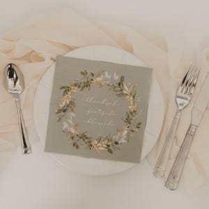 Puede incluir: Una servilleta cuadrada verde salvia con un diseño de corona floral y las palabras "thankful, grateful, blessed" en escritura blanca. La servilleta está sobre un plato blanco, con una cuchara, un tenedor y un cuchillo.