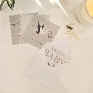 Könnte beinhalten: Ein Set aus Karten mit gewelltem Rand, die eine Storchenillustration mit dem Text Welcome baby zeigen, sowie eine Karte in einem weißen Umschlag mit dem Text SWEET BABY in brauner Schrift, ergänzt durch goldfarbene Metallbüroklammern und ein Glaswindlicht.