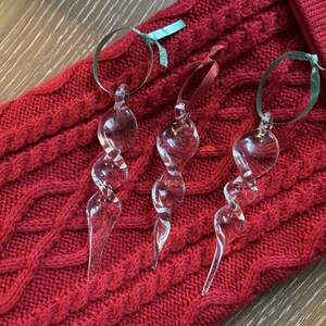 Puede incluir: Tres adornos de vidrio transparente en forma de espiral. Los adornos cuelgan de una cinta roja y verde sobre una superficie de punto rojo.