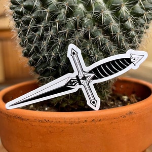 May include: A black and white sticker of a dagger with a cross-shaped hilt, placed in front of a cactus in a terracotta pot. The dagger design is detailed with a blade and hilt. The cactus is green and spiky.