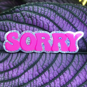Könnte beinhalten: Rosa glitzernder Aufkleber mit dem Wort "SORRY" in Blasenschrift.