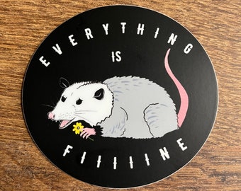 Puede incluir: Una pegatina redonda negra con una ilustración de un dibujo animado de zarigüeya. La zarigüeya tiene pelaje gris, una cola rosa y sostiene una flor amarilla. El texto blanco alrededor del borde dice "EVERYTHING IS FINE".