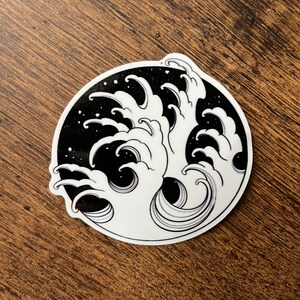 以下が含まれることがあります： 波が砕ける様子をスタイリッシュに描いた白黒のステッカー。波は伝統的な日本のスタイルで描かれており、渦巻くダイナミックなデザインが特徴です。