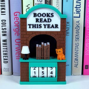 Può includere: Un contatore di libri verde acqua e marrone con la scritta "BOOKS READ THIS YEAR". Il contatore ha un design a camino in mattoni, una piccola statuina di gatto arancione e una lampada in miniatura. Il contatore mostra il numero 154.