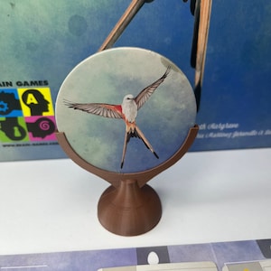 May include: A brown wooden stand with a round top holds a circular disc with a watercolor painting of a white-tailed swallow flying against a blue and green watercolor background.