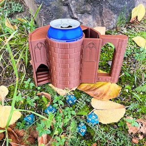 Op de afbeelding: Een bruine 3D-geprinte kasteelvormige blikjeshouder met een blauwe blikje erin. De houder heeft een kleine deur en ramen. Er zijn verschillende blauwe dobbelstenen rond de houder verspreid op een bed van groen mos en bruine bladeren.