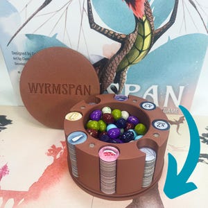 Könnte beinhalten: Ein brauner Holzspielorganizer mit den Worten "Wyrmspawn" auf dem Deckel. Der Organizer hat ein kreisförmiges Fach, das mit bunten Spielsteinen gefüllt ist. Der Deckel ist geöffnet und der Organizer steht auf einem Tisch mit einer Spielbox im Hintergrund.