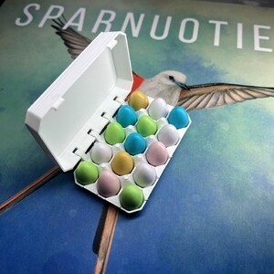 May include: A white cardboard egg carton with a lid open, revealing a variety of pastel coloured candy eggs. The carton is sitting on a blue and green background with a bird illustration.