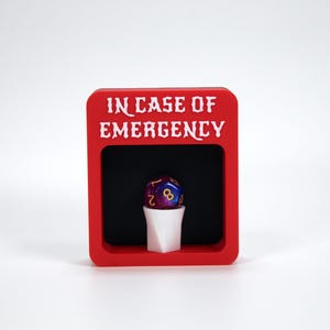 Könnte beinhalten: Eine rote Box mit dem Text "IN CASE OF EMERGENCY" in weißen Buchstaben. In der Box befindet sich ein weißer Becher mit einem lila und blauen Würfel mit goldenen Zahlen.