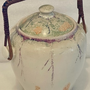 Może przedstawiać: Biały ceramiczny słoik z pokrywką i plecionym uchwytem. Pokrywka ma zielono-pomarańczowy kwiatowy wzór, a słoik fioletowe i pomarańczowe akcenty. Uchwyt wykonany jest z brązowego, plecionego materiału.