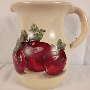 Puede incluir: Una jarra de cerámica de color crema con una ilustración pintada de tres manzanas rojas brillantes con hojas verdes en la parte frontal. La jarra tiene un cuerpo ancho y redondeado, un asa curva en el lado derecho y un pequeño pico vertedor.