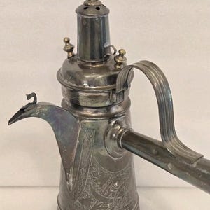 May include: Antique silver-colored metal coffee pot with a long spout and handle. The pot features intricate engraved designs and a bird-shaped finial on the lid. The pot has a decorative handle and spout, and is likely used for serving coffee or tea.