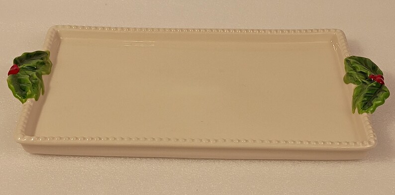 May include: An off-white ceramic serving tray, rectangular in shape, with a beaded edge. It has green holly leaf handles with red berries. The tray is approximately 30 cm long and 20 cm wide, ideal for festive occasions.