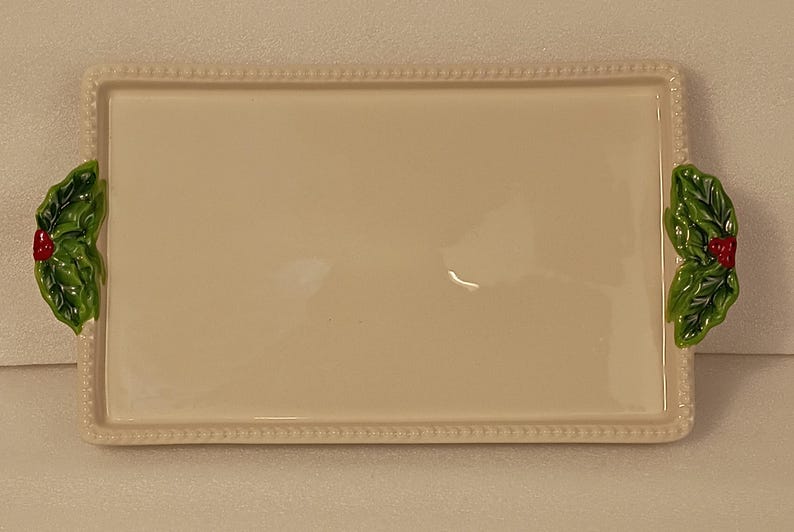 May include: Cream-coloured rectangular ceramic serving tray with a beaded edge. It has green holly leaf and red berry accents on each end. A decorative piece for serving food, perfect for festive occasions.