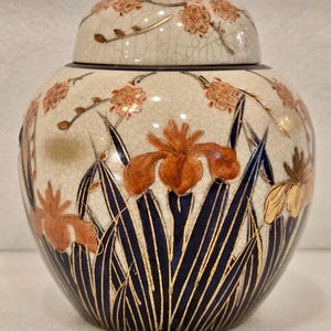 May include: A ceramic ginger jar with a lid, featuring a cream-colored background with a crackle glaze. The jar is adorned with orange iris flowers, gold accents, and dark blue stylized leaves. The lid is decorated with orange floral patterns.