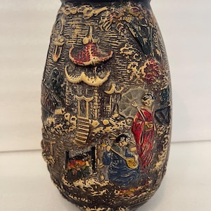 May include: A brown ceramic vase with a detailed relief design depicting an Asian landscape with figures, pagodas, and trees. The vase has a dark brown rim and a textured surface, showcasing a vintage aesthetic.