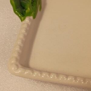 May include: An off-white ceramic serving platter with a decorative border. The top left corner features a green holly leaf design with a red ladybug accent. The border has a raised, textured pattern.