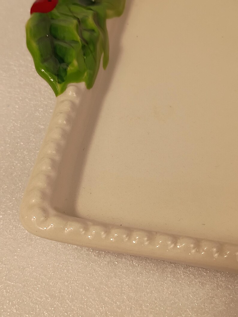 May include: An off-white ceramic serving platter with a decorative border. The top left corner features a green holly leaf design with a red ladybug accent. The border has a raised, textured pattern.