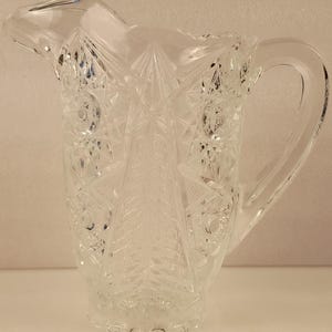 May include: Clear glass pitcher with a decorative, cut-glass pattern. The pitcher features a handle and a spout for pouring. The design includes starburst and geometric shapes, creating a vintage aesthetic. The pitcher is likely used for serving beverages.