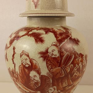 May include: A cream-colored ceramic ginger jar with a lid, featuring a detailed illustration in shades of red. The artwork depicts several figures in a landscape setting. The jar has a crackle glaze finish.