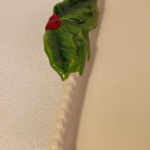 May include: An off-white ceramic serving tray, rectangular in shape, with a decorative holly leaf and red berry detail. The holly is green and glossy, providing a festive accent to the tray's design.