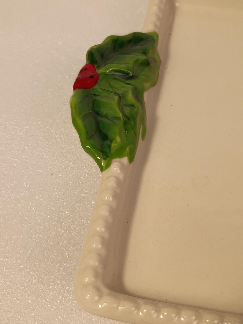 May include: An off-white ceramic serving tray, rectangular in shape, with a decorative holly leaf and red berry detail. The holly is green and glossy, providing a festive accent to the tray's design.