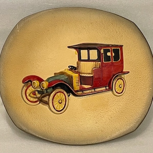 May include: A round, beige decorative plate featuring a raised relief of a vintage red car with yellow wheels. The car has a black roof and a yellow interior. The plate has a textured surface and a slightly irregular edge.