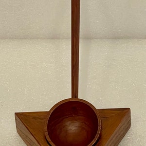 Puede incluir: Un soporte triangular de madera con un pequeño cuenco de madera y una larga clavija de madera. El soporte está hecho de madera oscura con veta natural.