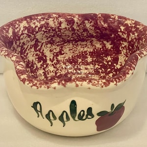 May include: A white ceramic bowl with a pink and white speckled glaze. The bowl has a scalloped rim and says "Apples" in green lettering with a green apple illustration.