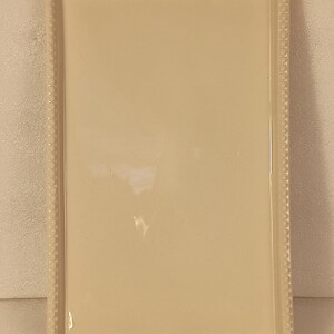 May include: Cream-coloured rectangular ceramic serving tray with a beaded edge. The tray is adorned with green holly leaves and red berries at the top and bottom, suggesting a festive theme. Suitable for serving food.