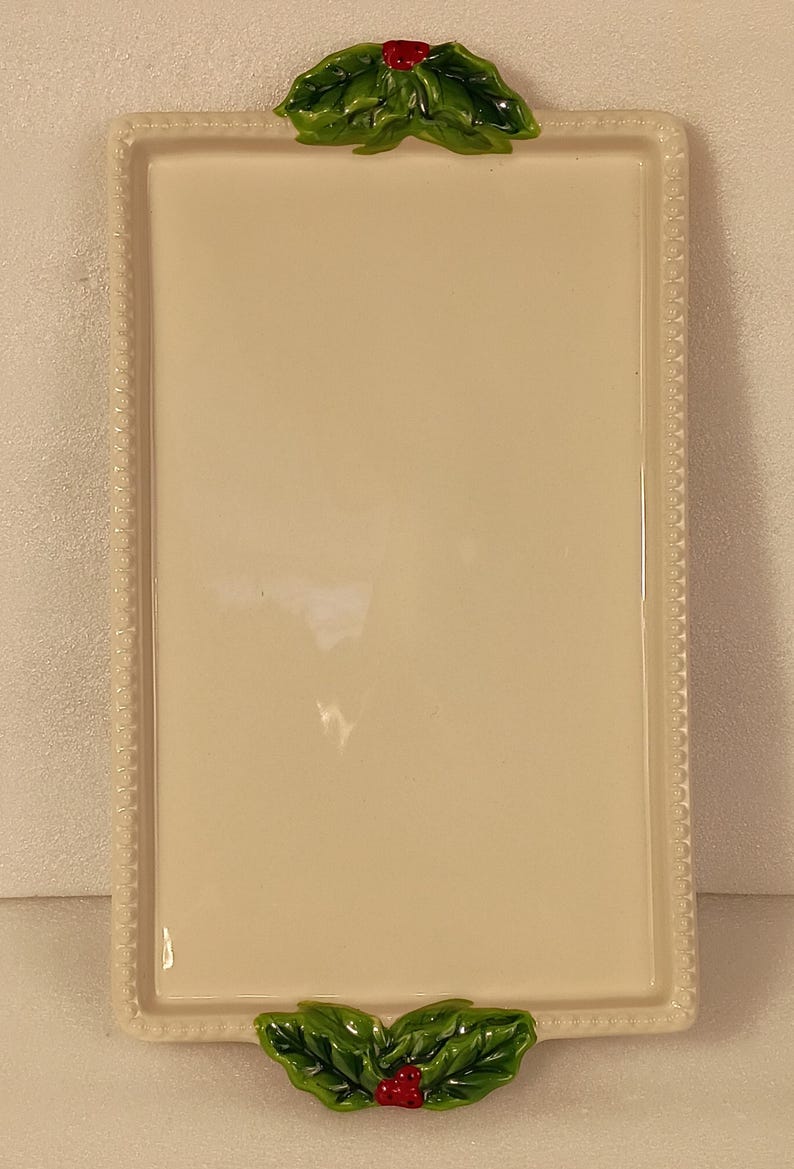 May include: Cream-coloured rectangular ceramic serving tray with a beaded edge. The tray is adorned with green holly leaves and red berries at the top and bottom, suggesting a festive theme. Suitable for serving food.