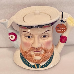 May include: A white ceramic teapot shaped like a man's head with a beard and mustache. The teapot has a lid and a spout. The teapot is decorated with red and white flowers and the words "James Sadler".