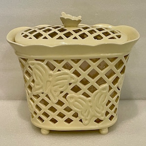 May include: A white ceramic container with a lid, featuring a lattice design and butterfly cutouts. The container has a scalloped rim and sits on three small feet.