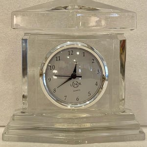 Puede incluir: Un reloj de cristal transparente con una esfera plateada y agujas negras. El reloj tiene la forma de un pequeño edificio con una base cuadrada y una parte superior en forma de pirámide. La esfera del reloj tiene números romanos negros y las palabras "LENOX QUARTZ" impresas en ella.