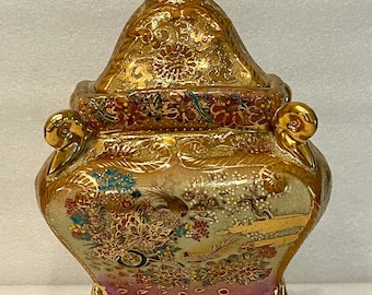 May include: An ornate, gold-coloured ceramic container with a lid. The container features a detailed floral and landscape design with red, blue, and green accents. It has three small feet and a handle on the lid.