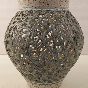 Może przedstawiać: Ceramiczny wazon o unikalnym ażurowym wzorze. Wazon ma jasnoniebieską i szarą kolorystykę z teksturowaną powierzchnią. Złożony wzór przypomina liście, tworząc dekoracyjny i artystyczny wygląd. Wazon ma około 25 cm wysokości.