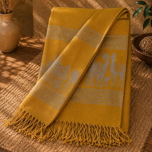 May include: A mustard yellow throw blanket featuring a repeating grey geometric Greek key border and a central pattern of grey camel silhouettes. The rectangular textile has a soft texture and is finished with long, knotted fringe along the edges.