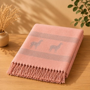 Puede incluir: Una manta de color rosa suave con un patrón de borde geométrico gris repetitivo y siluetas de llamas grises. La manta está terminada con largos flecos de color rosa a juego a lo largo del borde inferior, creando un aspecto texturizado.