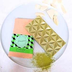 May include: A green matcha white chocolate bar with a geometric pattern on a white plate with a white background. The bar is wrapped in a brown paper wrapper with a green and black label that says "Raw, Organic, Rawsome, Vegan Chocolate, Matcha, Sesame White Chocolate".