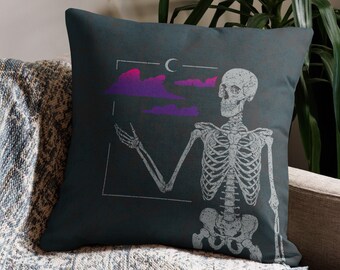 May include: A dark blue throw pillow with a skeleton holding a purple and pink cloud in front of a crescent moon. The skeleton is white and the background is dark blue.