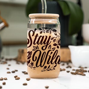 Puede incluir: Un tarro de vidrio con tapa de bambú y una pajita de vidrio. El tarro está lleno de un líquido marrón claro y tiene un gráfico en blanco y negro que dice "Stay Wild" con diseños florales. El tarro está sentado sobre una superficie blanca con granos de café esparcidos a su alrededor.