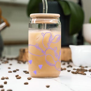 Puede incluir: Un tarro de vidrio con tapa de madera y un diseño de cactus morado lleno de café helado y una pajita de vidrio transparente.