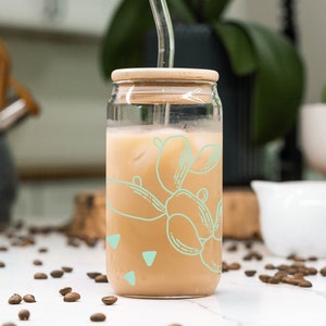 Puede incluir: Un tarro de vidrio con tapa de bambú y una pajita transparente llena de café helado. El tarro tiene un fondo marrón claro con un diseño abstracto verde y blanco.
