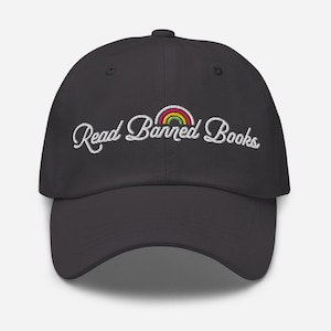 May include: A dark gray baseball cap with the embroidered text "Read Banned Books" in white script. A small rainbow is arched above the text. The cap has a curved brim and a button on top.