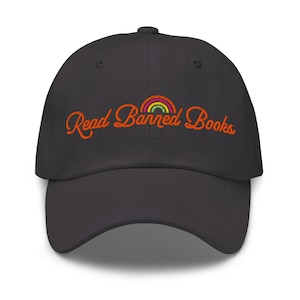 Peut inclure: Casquette de baseball gris foncé avec l'inscription brodée "Read Banned Books" en orange, surmontée d'un petit arc-en-ciel. L'arc-en-ciel présente des rayures rouges, oranges, jaunes, vertes et violettes. La casquette a une visière incurvée.