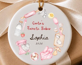 May include: A round white ceramic ornament featuring a gold hanging string and a watercolor illustration of baking tools including a pink mixer, rolling pin, and oven mitts. The text reads Santa's Favorite Baker, Sophia, and 2025.