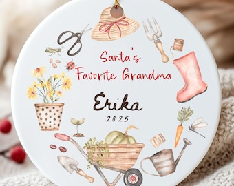 May include: A white ceramic Christmas ornament with a gold string. The ornament features a watercolor design of gardening tools, a straw hat, and the text "Santa's Favorite Grandma Erika 2025."