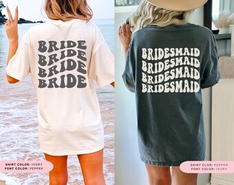 May include: Two t-shirts with a retro font design. The white shirt has the word "BRIDE" repeated four times in black. The gray shirt has the word "BRIDESMAID" repeated four times in white. The text on both shirts is in a wavy, retro font.