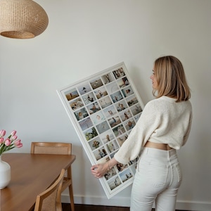 Op de afbeelding: Een grote, wit omkaderde fotocollage met talrijke kleine vierkante foto's. De collage wordt vastgehouden door een persoon in een cr&egrave;mekleurige trui en een witte broek. De achtergrond omvat een houten tafel, stoel en een geweven lamp.